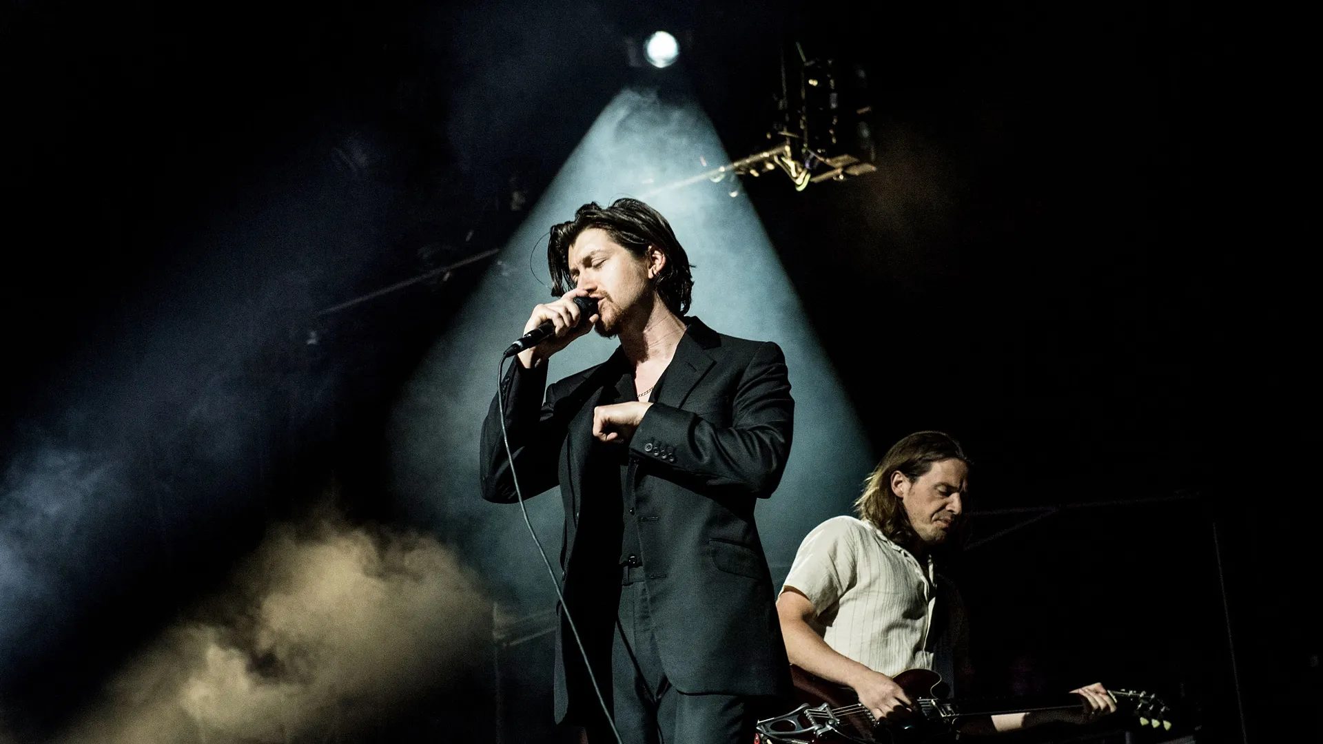 Vocalist in a dark suit singing into a microphone under a spotlight, with a guitarist behind